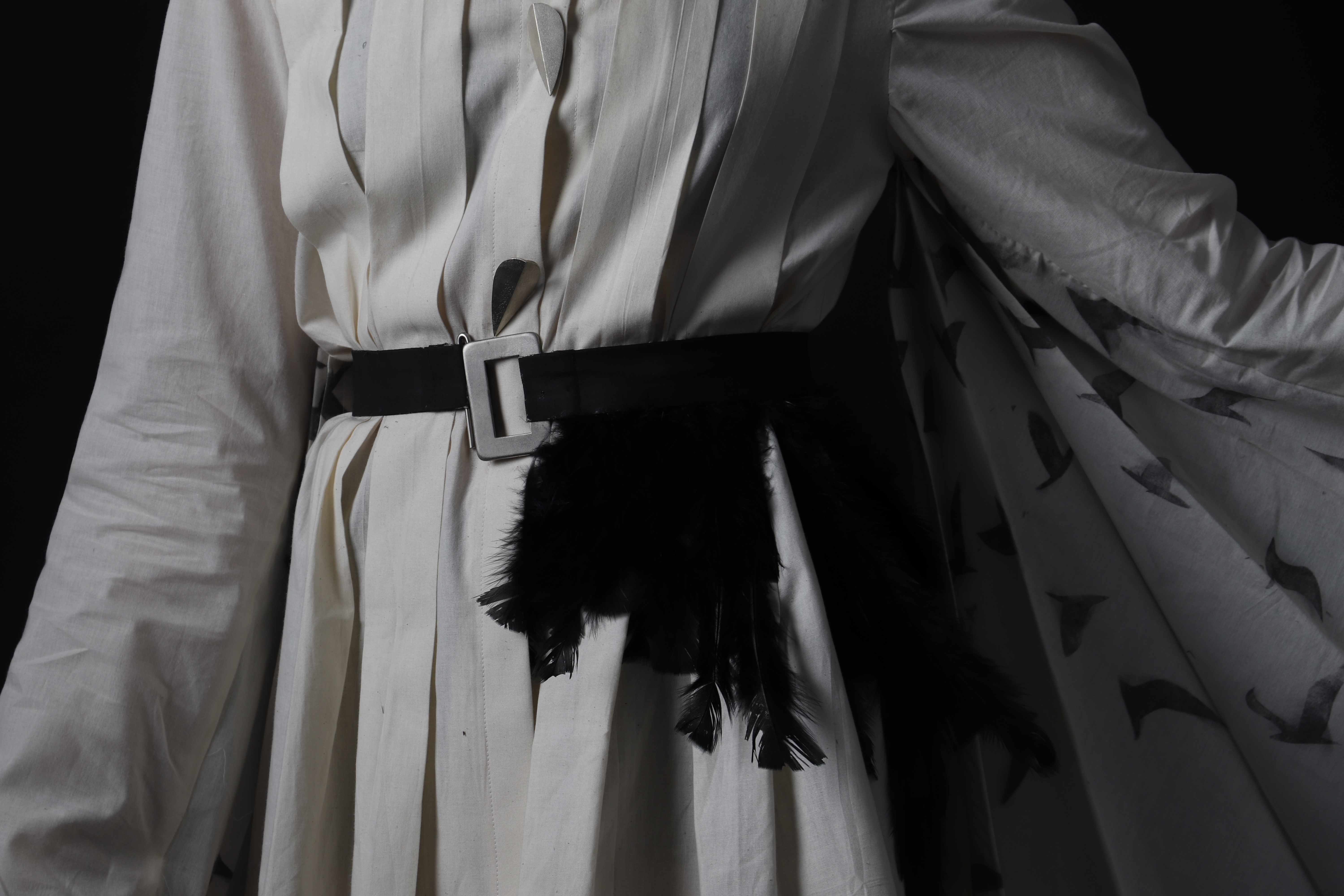 Photo rapprochée d’une chemise plissée en toile. Elle est tenue à la taille par une ceinture noire avec une boucle en argent et des plumes noires y sont attachées. Les manches sont longues et des oiseaux noirs sont imprimés dessus.