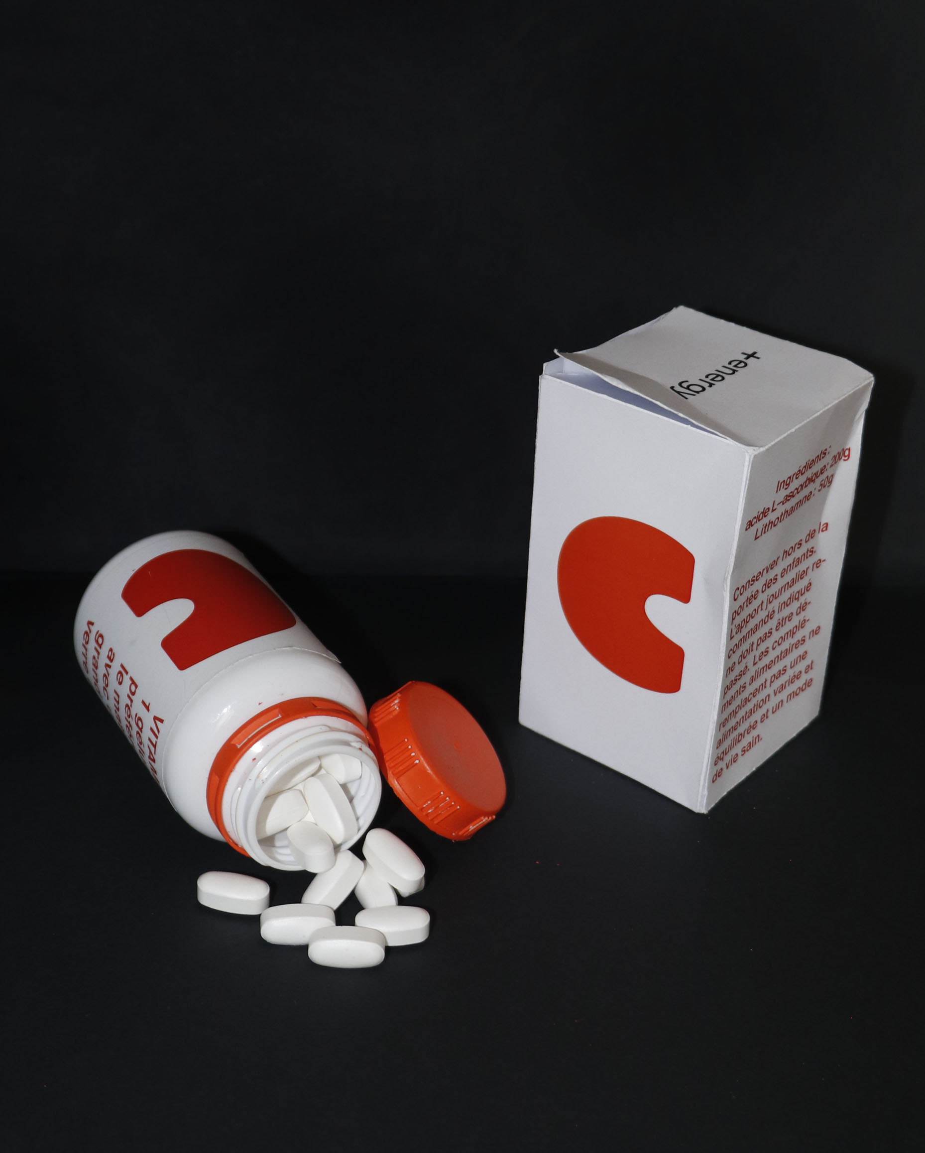 Photo d’une bouteille de vitamines ouverte et renversé. Les gélules blanches sont éparpillées. La bouteille est blanche avec un bouchon orange. Sur l’étiquette de la bouteille, on peut voir un gros « C » orange. À gauche de l’image, une boite en carton blanc reprend le design de l’étiquette.