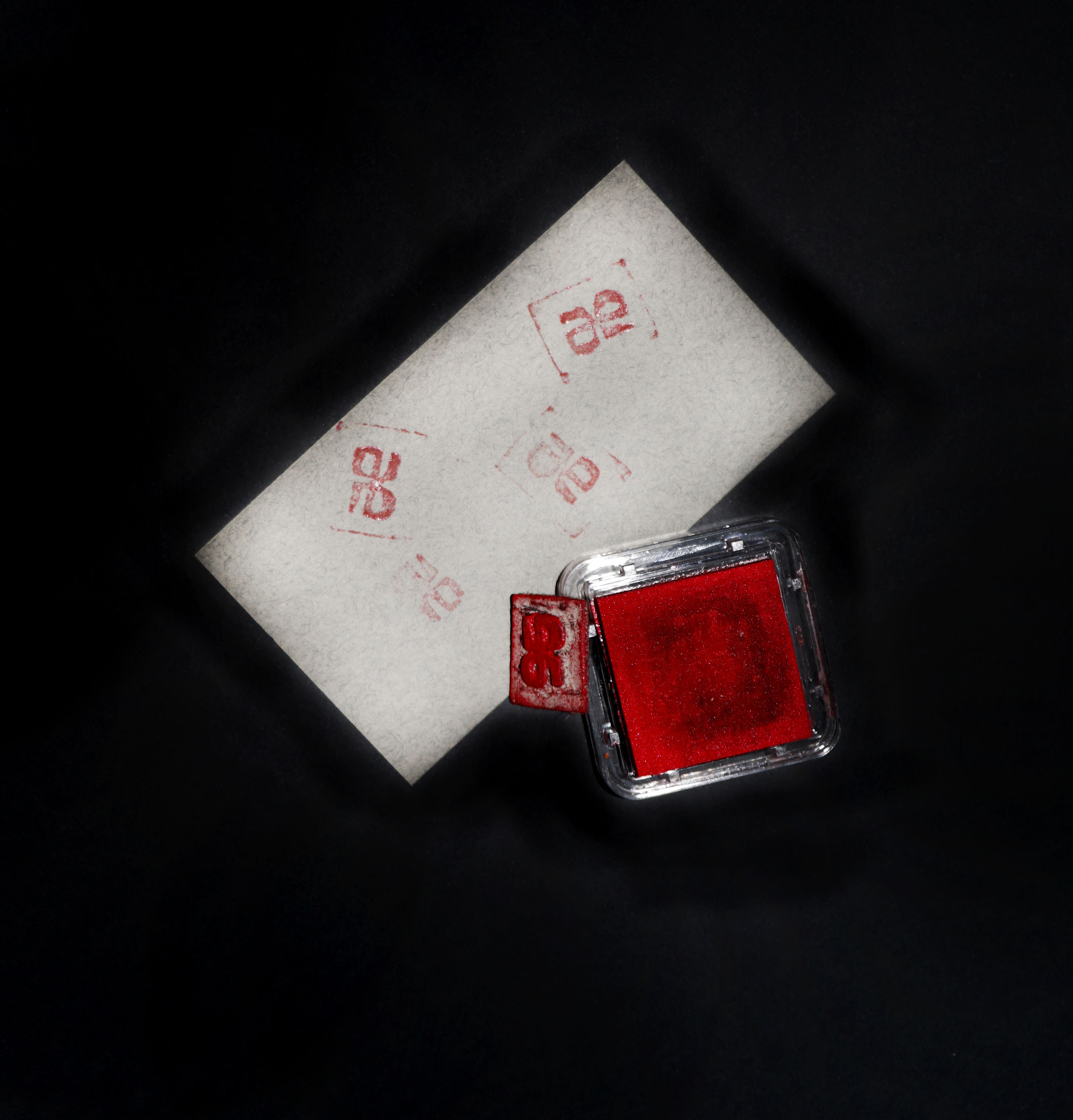 Photo d’une feuille grise tamponnée avec le sigle « JP » en rouge. À côté, le tampon est posé contre le coussin à encre.